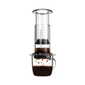 The Pull & Pour Shop AeroPress Clear coffee maker in transparent Tritan plastic brewing coffee into a glass mug.