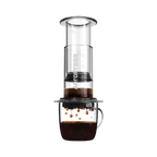 The Pull & Pour Shop AeroPress Clear coffee maker in transparent Tritan plastic brewing coffee into a glass mug.
