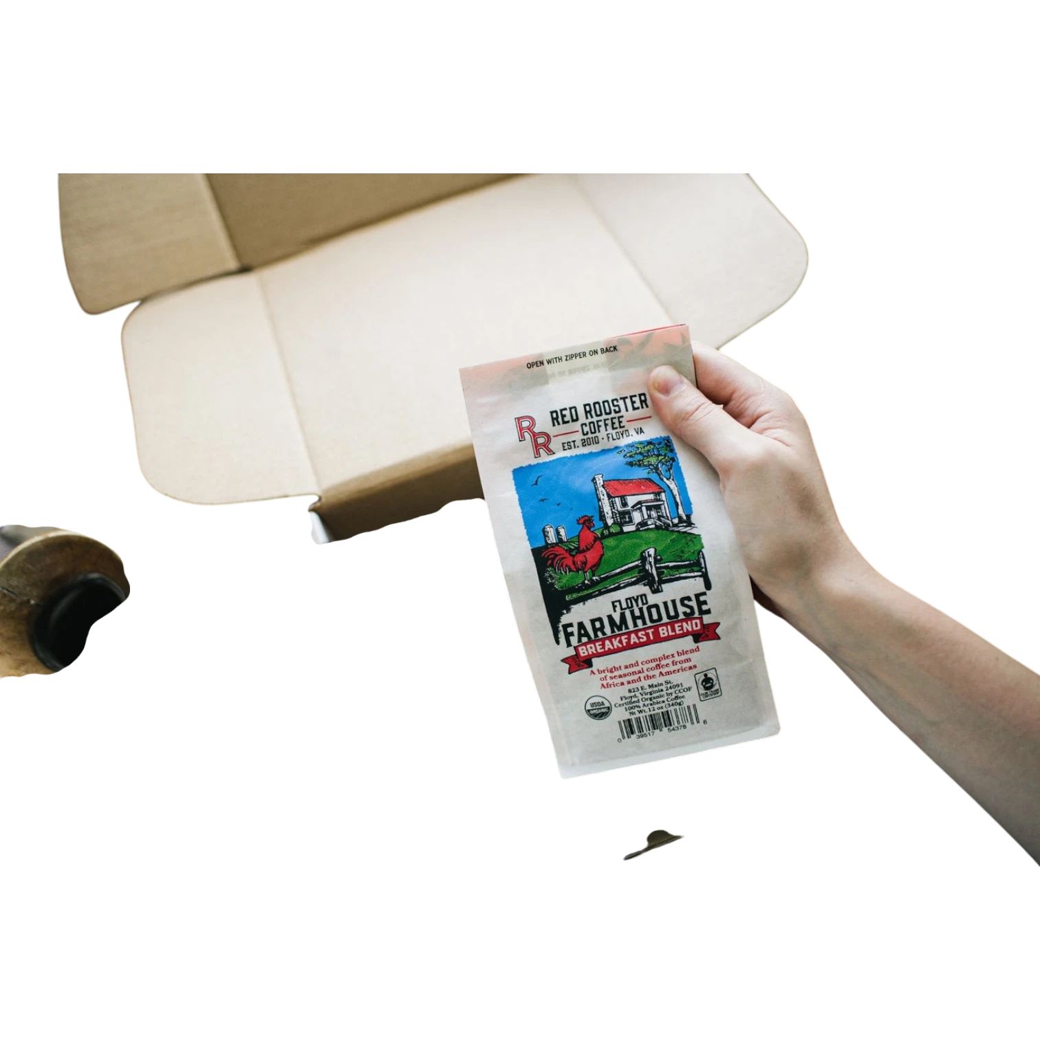 The Pull & Pour Shop hand holding a bag of Red Rooster Coffee Organic Floyd Farmhouse Breakfast Blend over a box